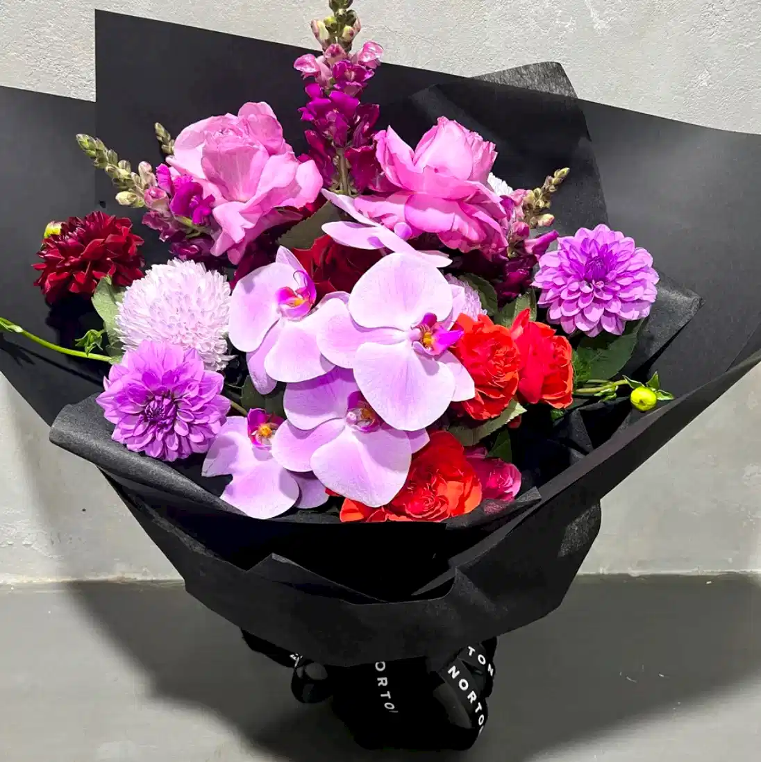 The Be Bold Seasonal Bouquet by FlowersonNortonSt showcases a vibrant mix of seasonal flowers, including pink phalaenopsis orchids, roses, dahlias, and snapdragons. This elegant arrangement is wrapped in black paper and presented on a gray surface.