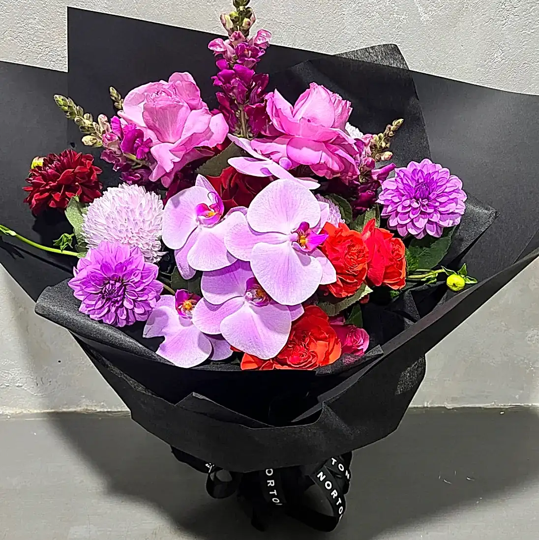 The Be Bold Seasonal Bouquet by FlowersonNortonSt showcases a vibrant mix of seasonal flowers, including pink phalaenopsis orchids, roses, dahlias, and snapdragons. This elegant arrangement is wrapped in black paper and presented on a gray surface.
