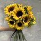 A hand holding a bouquet of "Sunny Sunflowers" from Flowers on Norton St shines against a textured gray wall, enhancing the radiant colors of the flowers' dark centers.