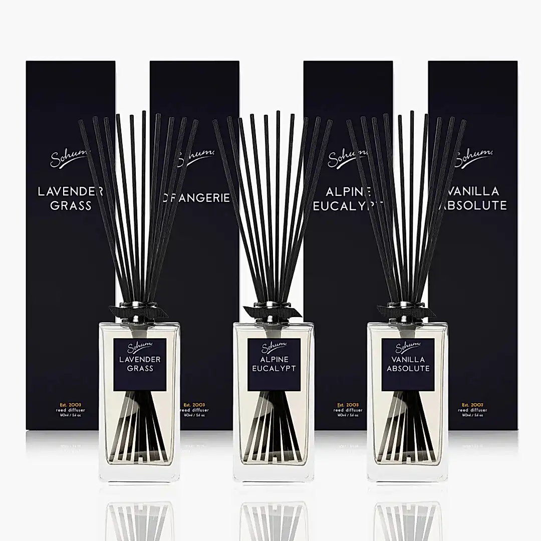 Three Sohum Fragrance Diffusers by Flowers on Norton St, featuring tall black sticks and matching containers, are displayed in front of dark boxes. The exquisite scents offered are Lavender Grass, Alpine Eucalypt, and Vanilla Absolute.