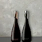 Two elegant, dark wine bottles against a textured gray background. Each bottle features a distinctive metallic accent on the neck—one gold, the other silver. Subtle text highlights their labels: "Flowers on Norton St Georg Jensen Sparkling Wine" and "Georg Jensen Mulled Wine.