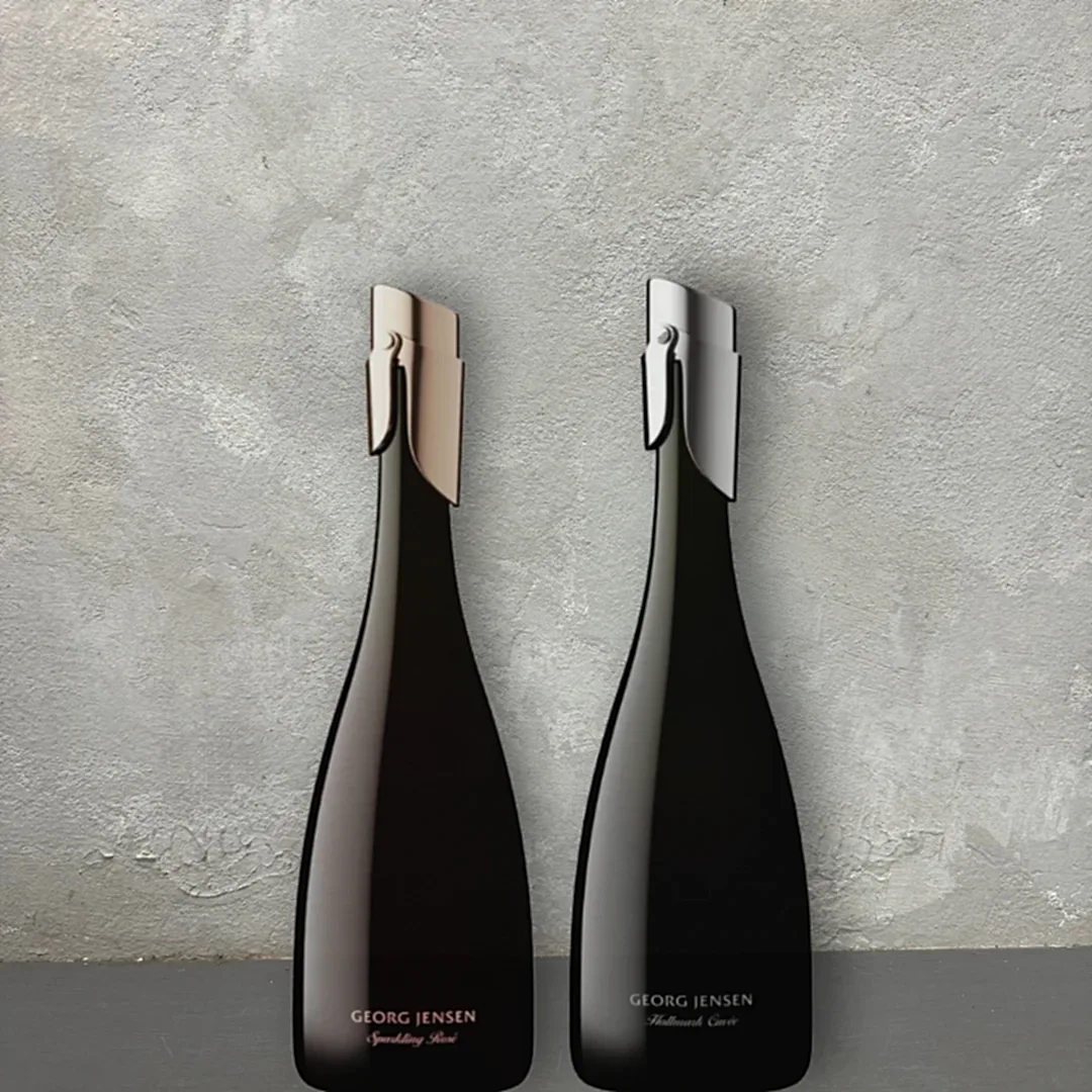 Two elegant, dark wine bottles against a textured gray background. Each bottle features a distinctive metallic accent on the neck—one gold, the other silver. Subtle text highlights their labels: "Flowers on Norton St Georg Jensen Sparkling Wine" and "Georg Jensen Mulled Wine.