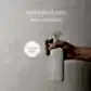 A hand holds a beige spray bottle labeled "AL.IVE BODY | Home & Kitchen" while text above reads "Flowers on Norton St - Kitchen Bench Spray Lemon & Pomegranate." This kitchen essential, featuring a circular "In Store Now" label against a light backdrop, brings freshness to your space.