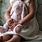 A child in a white dress gently holds the Lily & George Bunny by Flowers on Norton St, featuring floral-patterned ears and feet. The child's hands rest softly on the cuddly bunny, set against a subtle floral wallpaper backdrop.