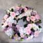 A Condolences Wreath by FlowersonNortonSt, showcasing pink roses, white hydrangeas, and purple eustomas with green leaves set against a light gray background.