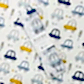 A delicate muslin wrap featuring a pastel print of blue, gray, and yellow cars, traffic lights, and triangles is ideal for swaddling babies. It is elegantly displayed on a coordinating background, complete with a label stating "Muslin Wraps by FlowersonNortonSt, 100% Cotton.