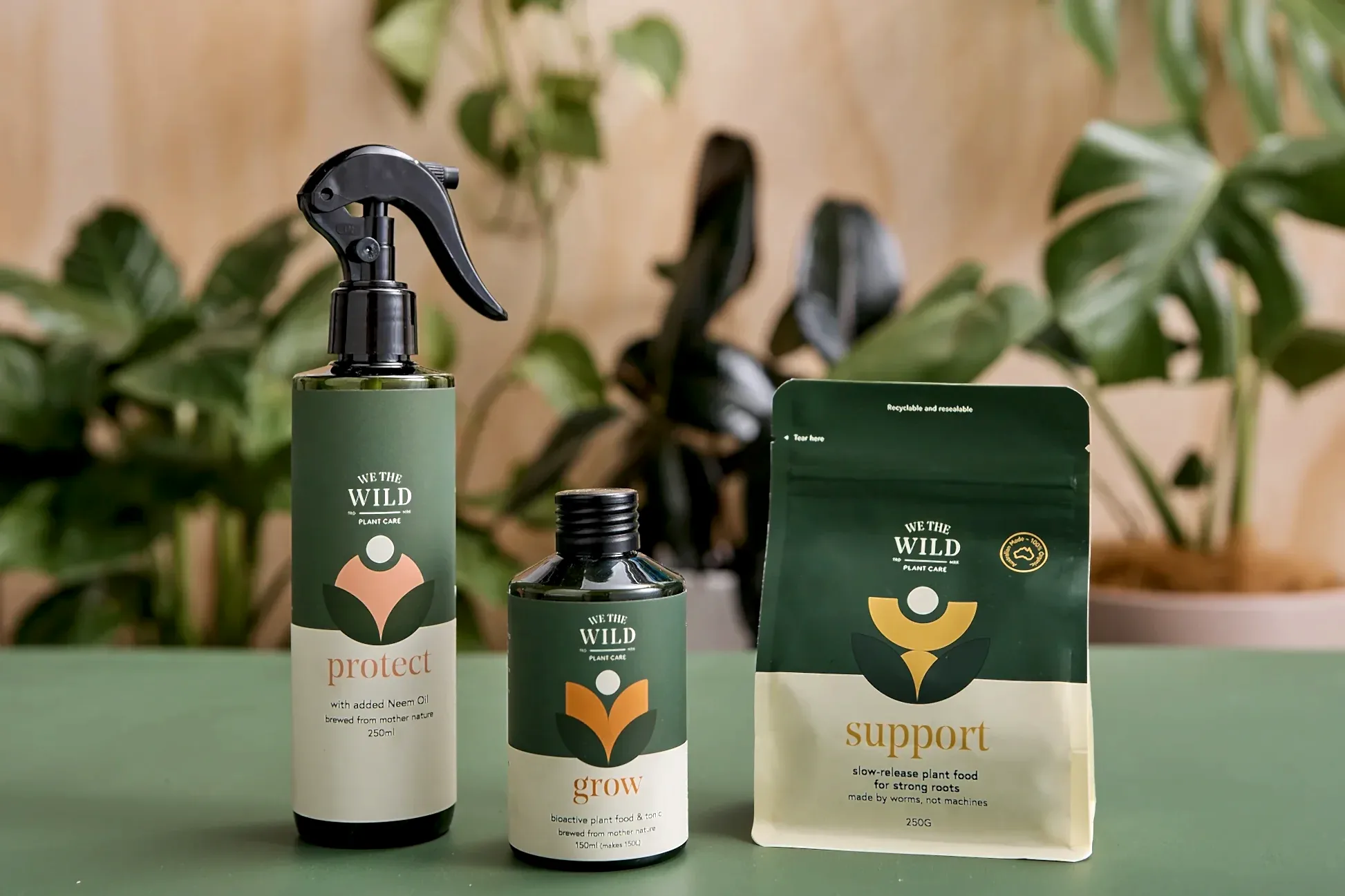 Presenting We The Wild Plant Care Products by FlowersonNortonSt: a collection of essential plant care items set against a verdant backdrop. This set features a spray bottle called "Protect," a smaller bottle titled "Grow," and a pouch named "Support." Each item boasts green and orange designs adorned with leaf illustrations, making them ideal for nurturing your indoor potted plants.