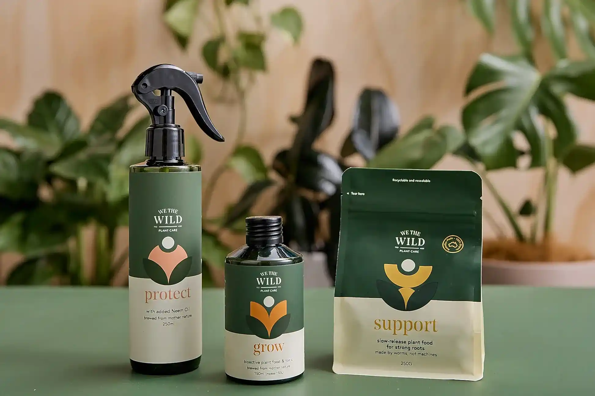 Displayed against a backdrop of plants are three organic plant care products from the "We The Wild" collection by FlowersonNortonSt. From left to right: a green and tan spray bottle labeled "Protect," designed for pest management; a small green and tan bottle labeled "Grow"; and a pouch labeled "Support.