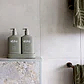 Two bottles of AL.IVE BODY | Earth Collection Duos from Flowers on Norton St, in olive green, sit on a small tray placed on a stone ledge in a minimalist bathroom. A matching green towel is draped over a wooden rod beside them against a light-colored tiled wall, showcasing their cruelty-free elegance.