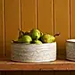 A Robert Gordon | Wheel Bowl from FlowersonNortonSt, filled with green pears, sits on a wooden shelf against a mustard-colored paneled wall.