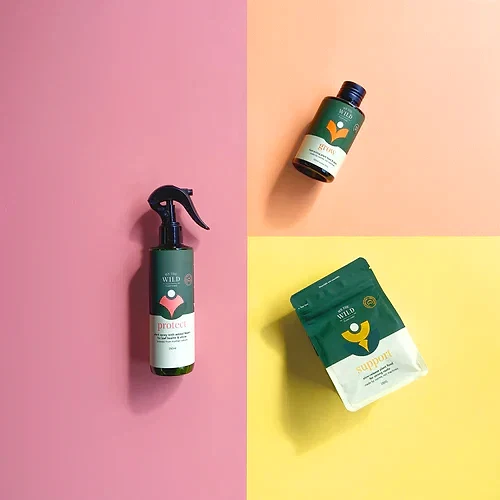 A flat lay photograph features the Plant Lovers Gift Set from FlowersonNortonSt, with three skincare products in green and white packaging that evoke a plant care theme, arranged against a vibrant pink and yellow background. On the left is a spray bottle that resembles pest spray, while the pouch is positioned at the bottom right and a bottle sits at the top right.