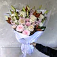 A hand holds the Serenity Sheath from FlowersonNortonSt, wrapped in white paper. This arrangement showcases a beautiful assortment of white lilies, pink roses, and white roses, complemented by additional pink and white blooms with green leaves on a light gray background—ideal as a sympathy tribute.