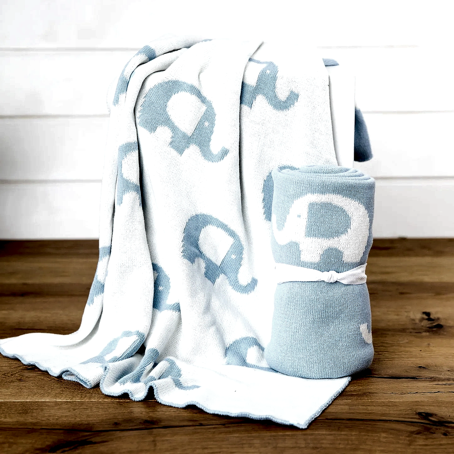 A cuddly Ellie Baby Blanket by FlowersonNortonSt, featuring a white base with blue elephants, drapes over a chair. It is accompanied by another Ellie Baby Blanket rolled up and tied with a white ribbon, both set gracefully on a wooden floor against a pristine white backdrop.