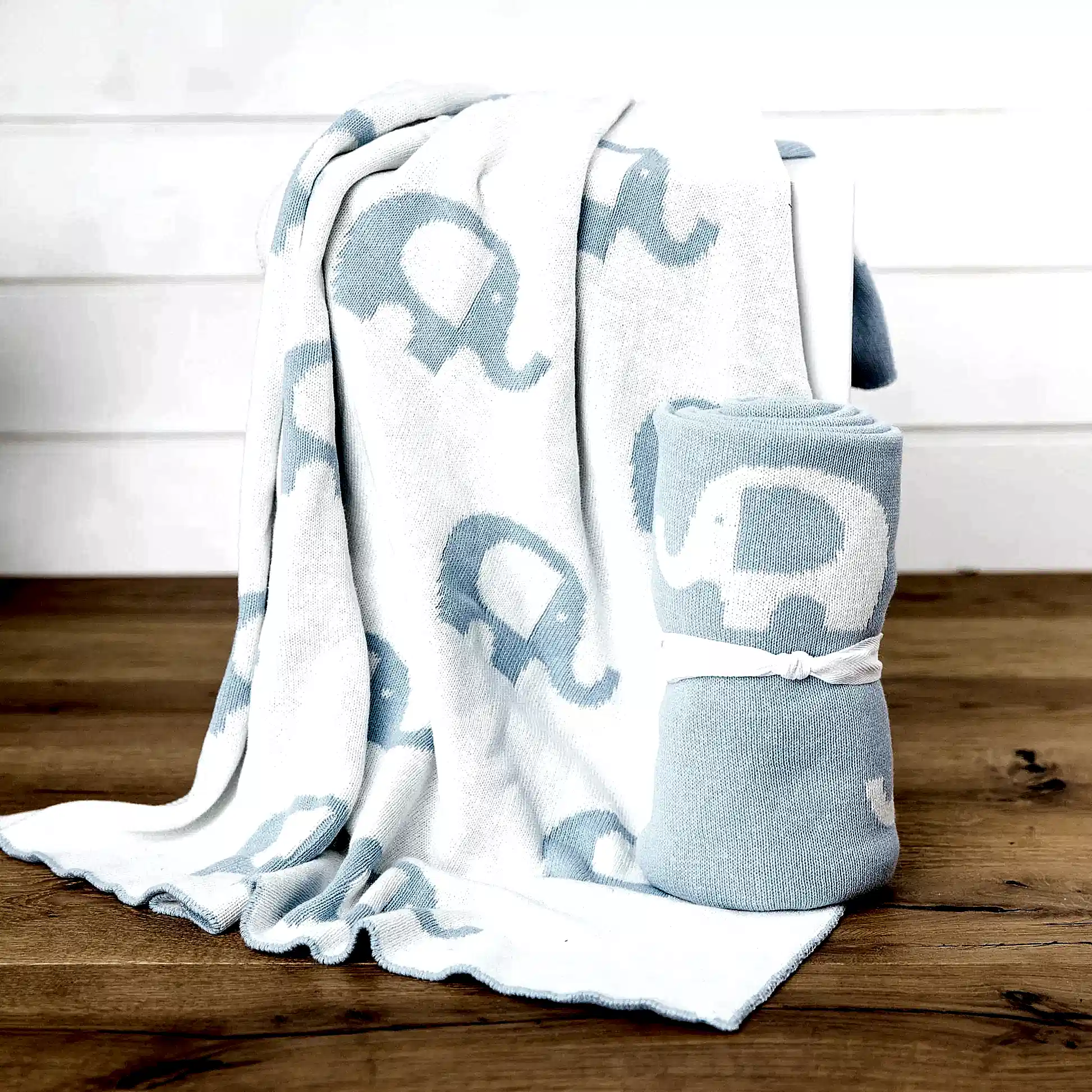 A cuddly Ellie Baby Blanket by FlowersonNortonSt, featuring a white base with blue elephants, drapes over a chair. It is accompanied by another Ellie Baby Blanket rolled up and tied with a white ribbon, both set gracefully on a wooden floor against a pristine white backdrop.