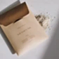 An elegantly designed beige envelope featuring "Flowers on Norton St" and "Body Scrub to Mask | Peggy Sue Co." rests gracefully on a white surface. Its flap is slightly open, exposing some of the white powder inside that promises to soften skin, while gentle shadows underscore its soothing benefits.