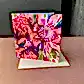 The FlowersonNortonSt Greeting Card boasts a stunning floral design with prominent pink, red, and green blooms. Perfect for any occasion, the card is shown partially open on a simple beige envelope against a brown surface and dark background.