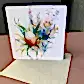 The FlowersonNortonSt Greeting Card showcases an eye-catching watercolor illustration of a vibrant floral bouquet with pastel green, yellow, and pink leaves and flowers. Ideal for any occasion, it rests on a brown surface alongside a beige envelope.