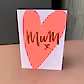 A beautifully designed greeting card from FlowersonNortonSt features a large pink heart with "Mum" elegantly scripted in gold, followed by a charming "x." Perfect for any occasion, this Greeting Card stands gracefully against a brown wooden background.