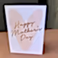 The FlowersonNortonSt Greeting Card is beautifully designed, featuring a large pink heart in the background with the words "Happy Mother's Day" elegantly scripted in gold. Perfect for any occasion, it rests gracefully on a brown surface against a wooden backdrop.
