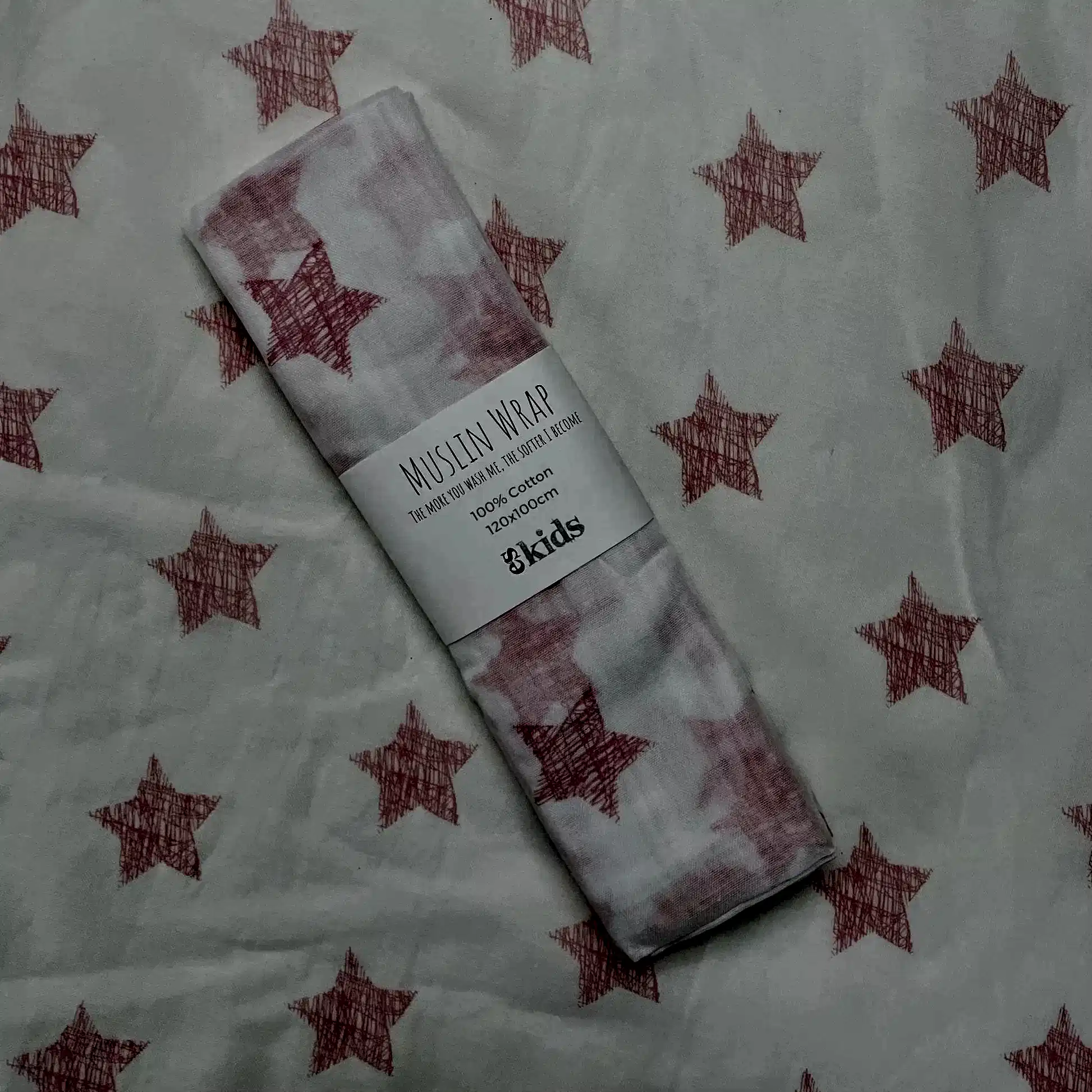 A Muslin Wrap from FlowersonNortonSt, featuring a charming pink star pattern, rests on matching fabric. The label indicates it's made of 100% cotton, ideal for swaddling and providing soft comfort.
