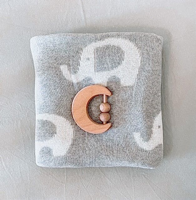 The Ellie Baby Blanket by FlowersonNortonSt, a soft and cuddly gray blanket adorned with charming white elephant patterns, is neatly folded. On top rests a crescent moon-shaped wooden teething toy featuring three beads, all set against a plain, light surface.