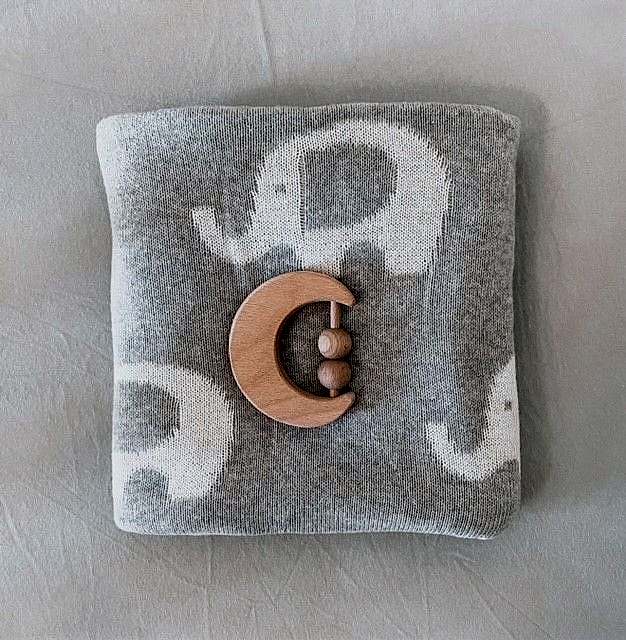 The Ellie Baby Blanket by FlowersonNortonSt, a soft and cuddly gray blanket adorned with charming white elephant patterns, is neatly folded. On top rests a crescent moon-shaped wooden teething toy featuring three beads, all set against a plain, light surface.
