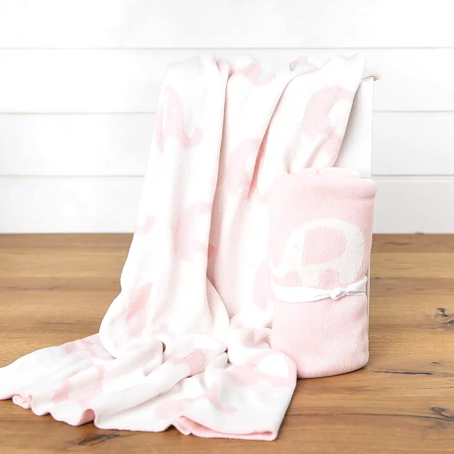 An Ellie Baby Blanket from FlowersonNortonSt, featuring a cuddly, soft white and pink elephant pattern, is draped over a white box. Another blanket with the same adorable design is rolled up, tied with a white ribbon, and placed beside it on a wooden surface.