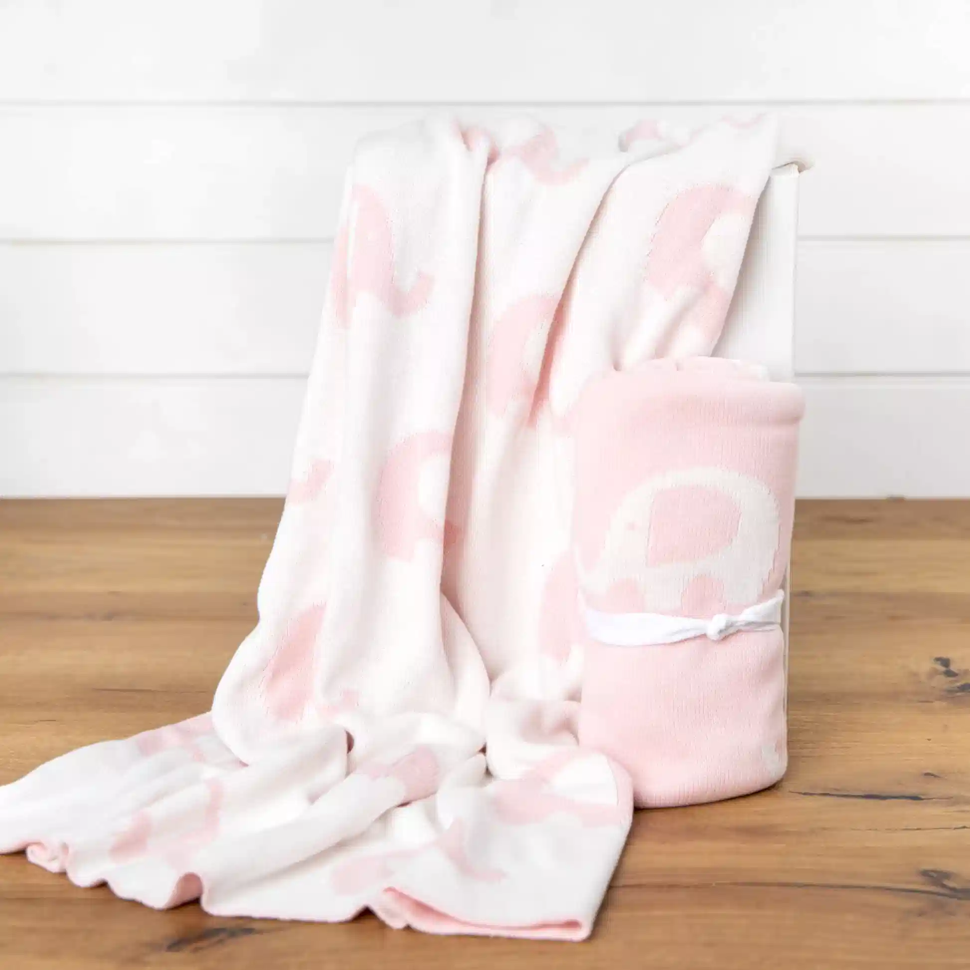An Ellie Baby Blanket from FlowersonNortonSt, featuring a cuddly, soft white and pink elephant pattern, is draped over a white box. Another blanket with the same adorable design is rolled up, tied with a white ribbon, and placed beside it on a wooden surface.