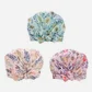 Three fashionable shower caps from Flowers on Norton St: one in light blue with green leaves, another with a white base featuring a pink and orange floral pattern, and a third in pastel pink embellished with red and blue flowers. Each cap is made from waterproof polyester and includes a gathered, knotted front.