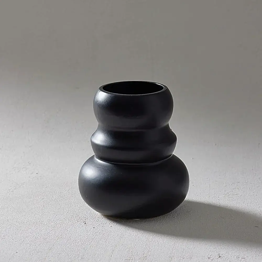 The Klein Vase, a handcrafted product by Flowers on Norton St, features a matte black finish and an unusual design with three distinct bulges. It rests on a light gray surface, casting a soft shadow to the right that accentuates its smooth texture and modern aesthetic reminiscent of organic forms.