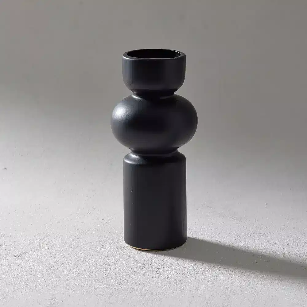A matte black ceramic vase from Flowers on Norton St, with an abstract and modern design inspired by a Klein Vase. It boasts a tall cylindrical base and a rounded, sculptural midsection, elegantly displayed against a plain light gray background with a subtle shadow.