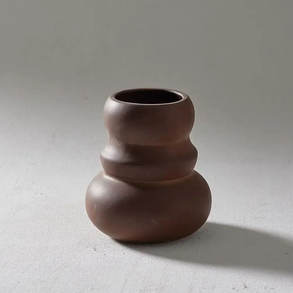 The Klein Vase by Flowers on Norton St rests on a smooth, light gray surface. With its brown, handcrafted ceramic body and a bulbous, tiered design reminiscent of organic forms, the vase boasts a matte finish and casts a gentle shadow to the right.