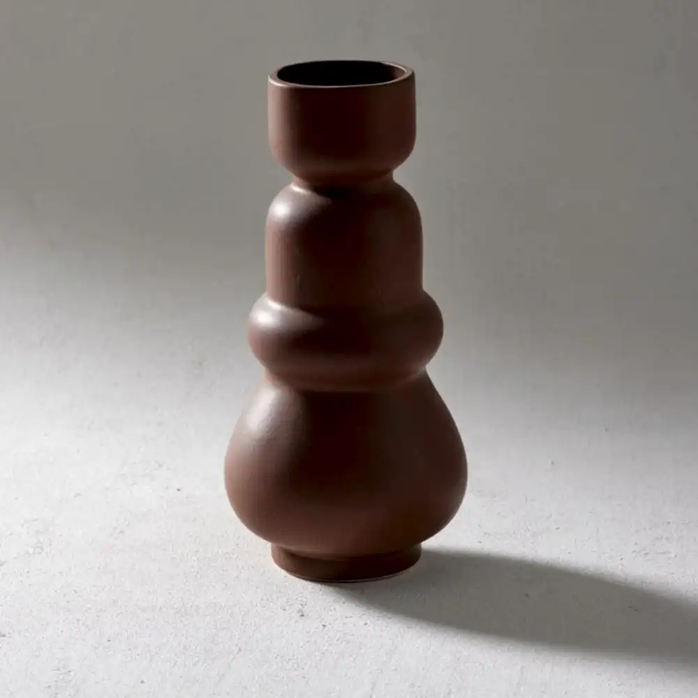 The Klein Vase, a creation by Flowers on Norton St, is a brown ceramic piece showcasing a modern, abstract design with rounded, stacked shapes that echo an organic form. This handcrafted vase stands on a light gray surface and casts a gentle shadow to the right.