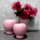 Flowers on Norton St's Pink Ceramic Vaseware elegantly showcases vibrant pink dahlias with lush green leaves. Beside them, two pink round ceramic vases rest on a gray surface, all set against a textured gray background, highlighting the exquisite beauty of modern vaseware.
