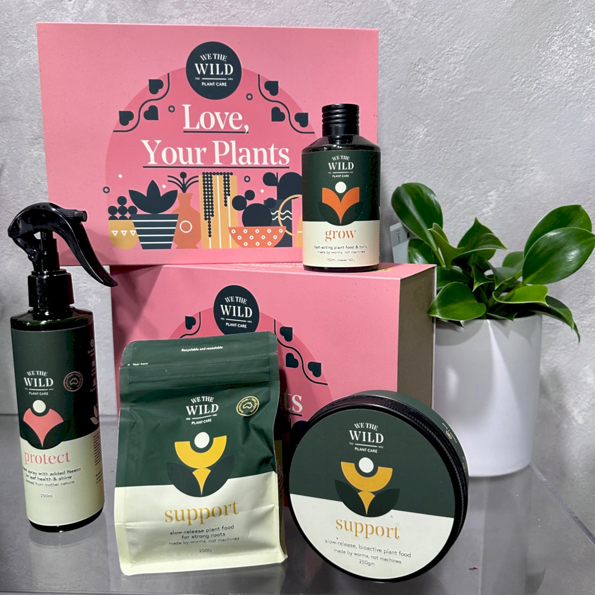 On the table, you can see a collection from the Plant Lovers Gift Set by FlowersonNortonSt. The items include a pest spray bottle, a growth concentrate called "grow," and both a pouch and tub labeled "support." Behind them is a pink sign adorned with plant illustrations that reads "Love, Your Plants.