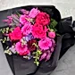 The Hot Pink Pop Mixed Flower Bouquet by Flowers on Norton St is a vibrant arrangement featuring bright pink and purple seasonal blooms, including roses and dahlias, all wrapped in black paper against a gray background.