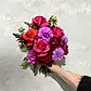 A hand holds a vibrant Be Bold Seasonal Bouquet by FlowersonNortonSt against a textured, light-colored wall. It features red roses, bright purple dahlias, magenta flowers, phalaenopsis orchids, and lush greenery.