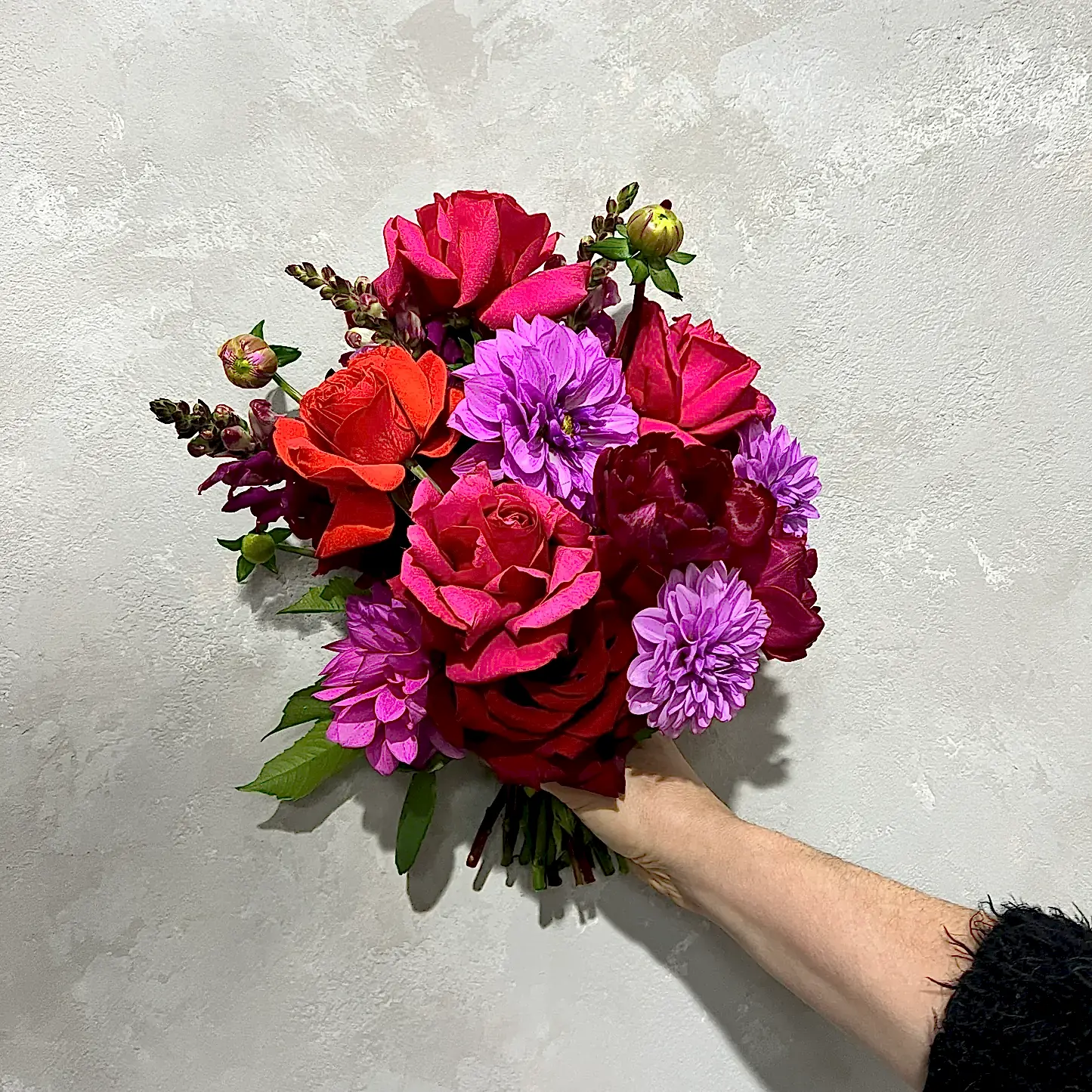 A hand holds a vibrant Be Bold Seasonal Bouquet by FlowersonNortonSt against a textured, light-colored wall. It features red roses, bright purple dahlias, magenta flowers, phalaenopsis orchids, and lush greenery.