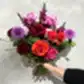 A hand holding the Be Bold Seasonal Bouquet by FlowersonNortonSt against a textured wall. This vibrant arrangement includes red, orange, and pink roses, purple dahlias, phalaenopsis orchids, along with other seasonal flowers for a lush and vivid display.