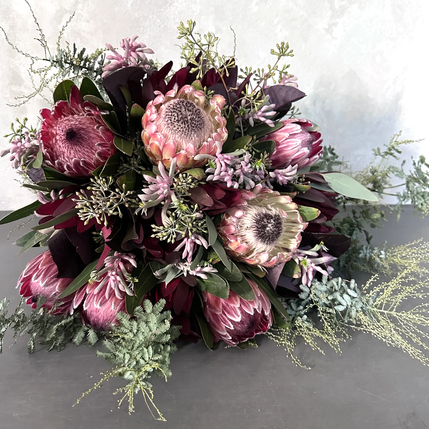 The Native Charm Bouquet by Flowers on Norton St features a luxe array of pink protea surrounded by dark purple and green foliage on a grey surface, with light green native sprigs enhancing its elegance against a neutral background.