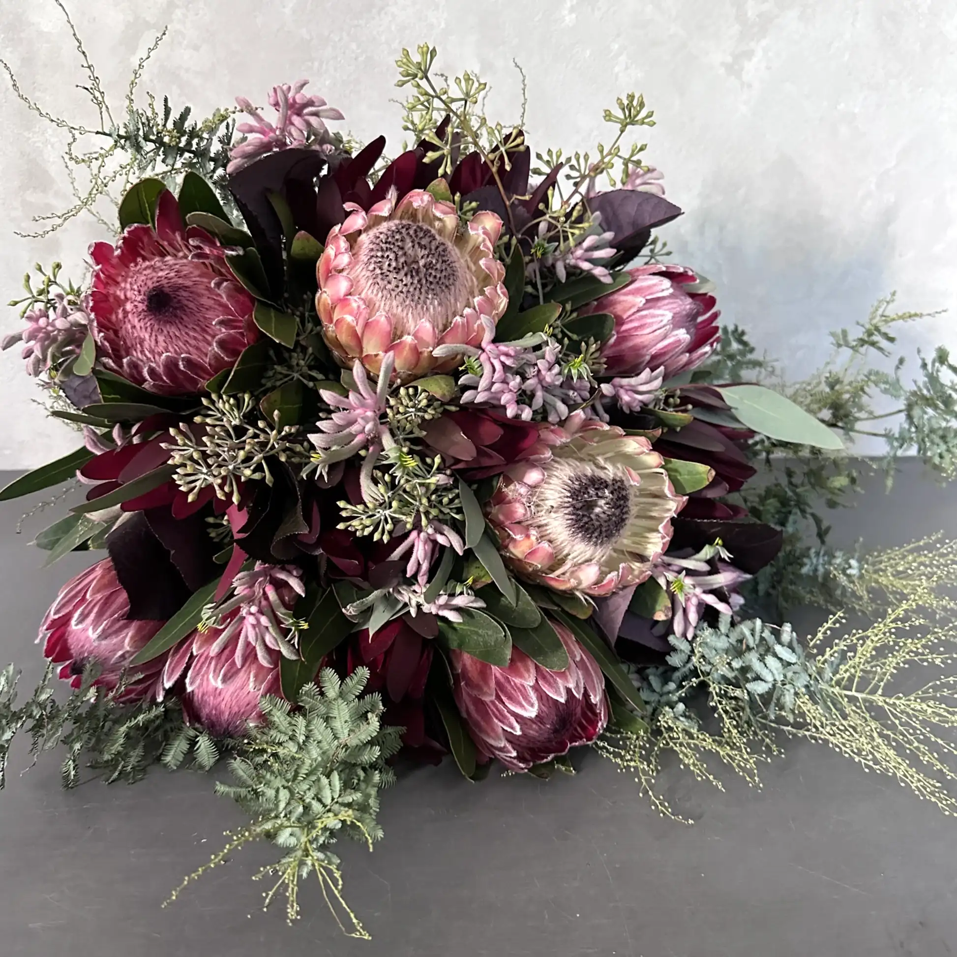 The Native Charm Bouquet by Flowers on Norton St features a luxe array of pink protea surrounded by dark purple and green foliage on a grey surface, with light green native sprigs enhancing its elegance against a neutral background.
