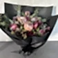 The Native Charm Bouquet by Flowers on Norton St is a striking arrangement wrapped in black paper, featuring large pink proteas, dark red flowers, and lush greenery. This luxurious bouquet stands out beautifully against a light gray backdrop.