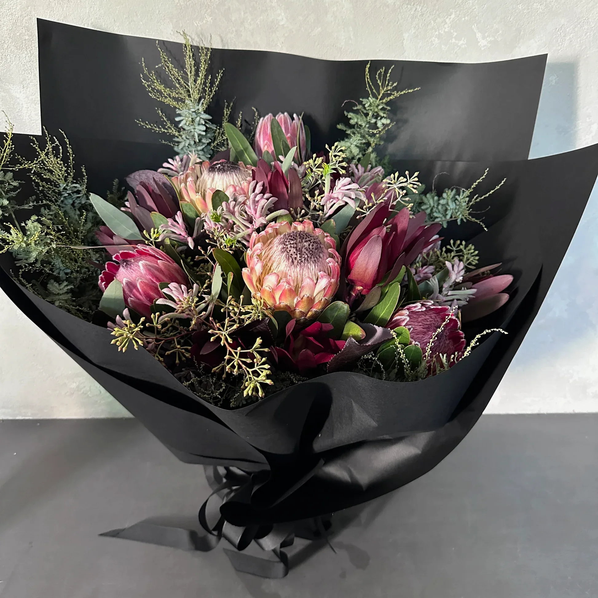 The Native Charm Bouquet by Flowers on Norton St is a striking arrangement wrapped in black paper, featuring large pink proteas, dark red flowers, and lush greenery. This luxurious bouquet stands out beautifully against a light gray backdrop.