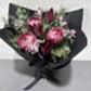 The "Little Native Bunch 2024" by Flowers on Norton St is a striking bouquet of pink protea, deep burgundy leaves, and delicate greenery, wrapped in black paper with a ribbon. This native flora arrangement is elegantly displayed on a gray surface against a light wall.