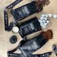 The Coffee Lover Gift Box by Flowers on Norton St features a flatlay presentation of three black coffee bags set against brown paper. The scene is accentuated with scattered coffee beans, a candle topped with a sleek black lid, white coffee capsules, hints of cocoa powder suggesting espresso chocolate notes, and a black ribbon adorned with white lettering.