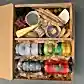 A gourmet gift box from Flowers on Norton St, known as the "Cans of Craft Beer Gift Box," includes three cans of craft beer, various cheeses, slices of cured meat, breadsticks, spiced nuts such as pistachios and almonds, crackers, grapes, a small jar of pesto, and a dessert cup—all elegantly arranged on a bed of wooden shavings to resemble a charcuterie board.