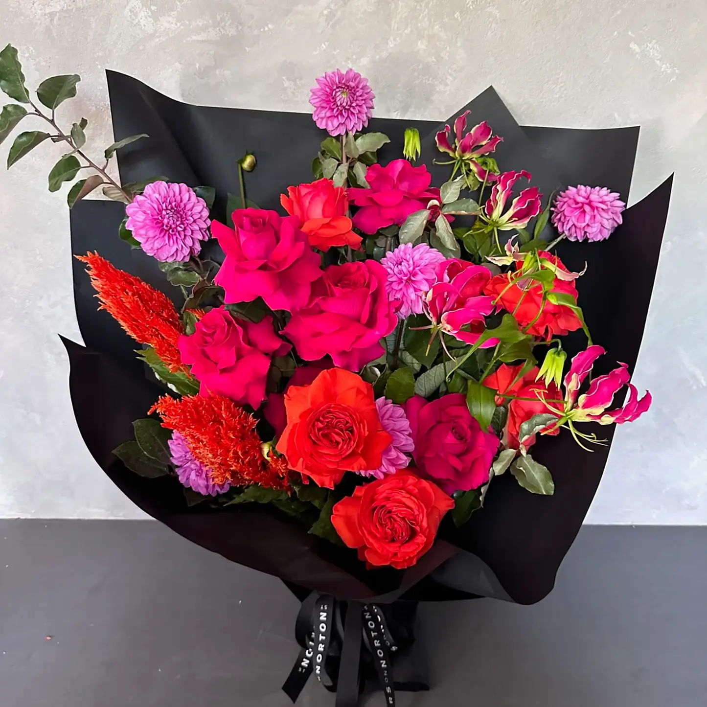 The Florist Choice - Red Nina Bouquet by FlowersonNortonSt features vibrant Red Nina and Colombian roses, bright pink blooms, and lush green foliage, elegantly wrapped in black paper with a black ribbon, set on a light gray backdrop.