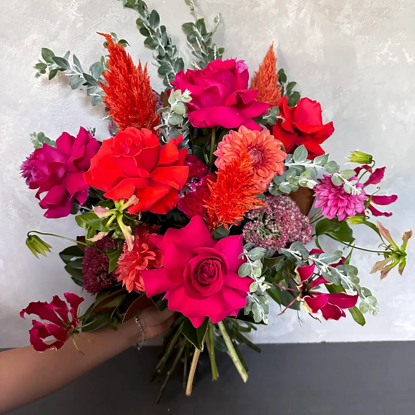 The Florist Choice - Red Nina Bouquet by FlowersonNortonSt showcases Red Nina roses with seasonal blooms in bright pink, red, orange, and purple hues, accented with eucalyptus leaves, all beautifully arranged against a light gray background.