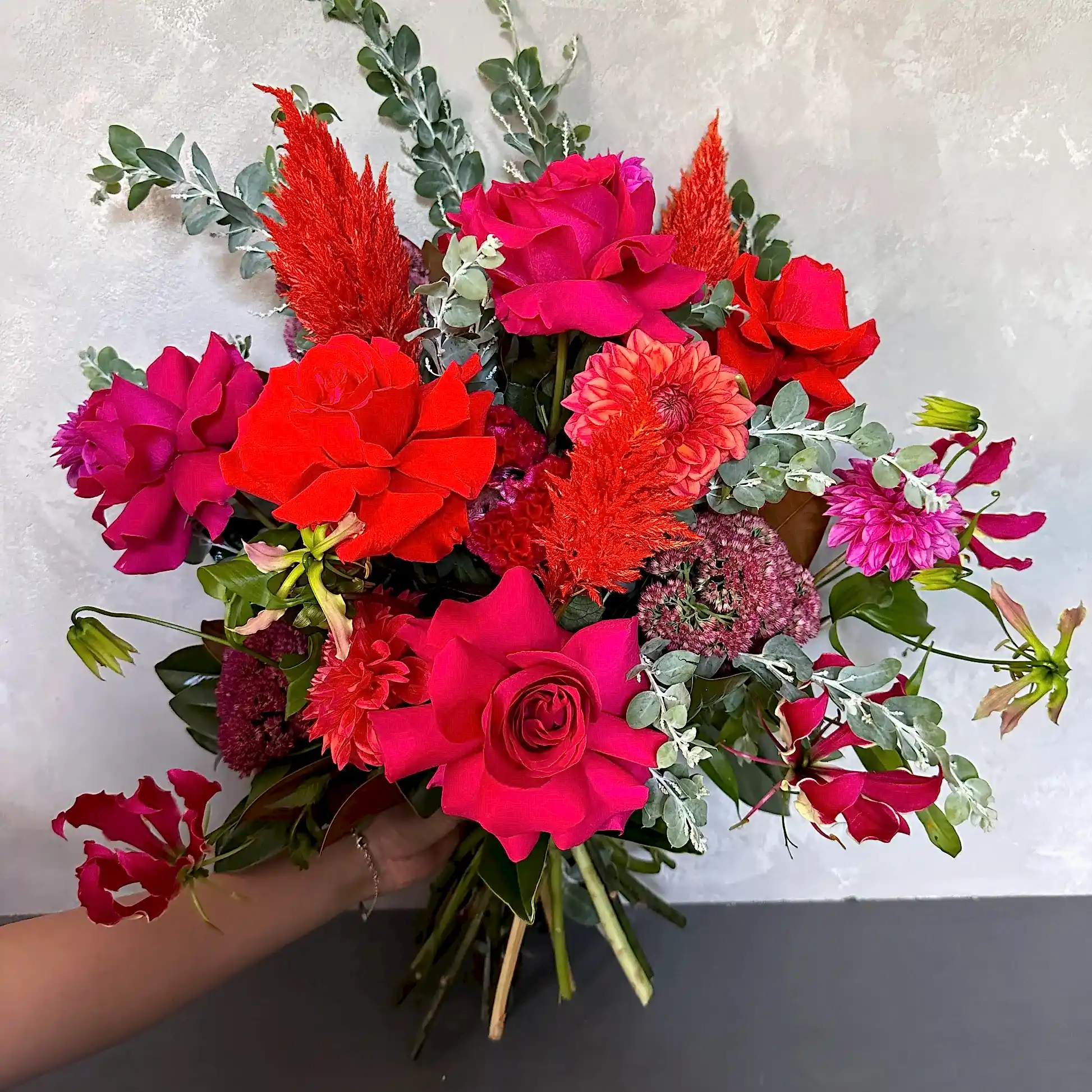 The Florist Choice - Red Nina Bouquet by FlowersonNortonSt showcases Red Nina roses with seasonal blooms in bright pink, red, orange, and purple hues, accented with eucalyptus leaves, all beautifully arranged against a light gray background.