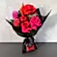 The Strawberry Kisses Mixed Bouquet 2024 by Flowers on Norton St, wrapped in black paper, features red and pink roses, dahlias, and a vibrant orange bloom. It includes a small package of Strawberry Kisses with a black ribbon against a light gray textured wall.
